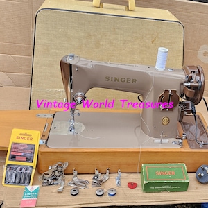 May include: A vintage Singer sewing machine in a tan colour, with a wooden base and accessories. The machine has the Singer logo on the arm. Accessories include a yellow Singer box, a green Singer box, and various attachments. A tan carrying case is in the background.