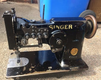 Puede incluir: Una máquina de coser Singer negra con una placa de identificación dorada y una gran rueda de metal. La máquina tiene un pedal de metal y una aguja.