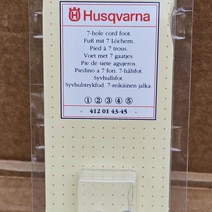 Könnte beinhalten: Ein Husqvarna 7-Loch-Kordelfuß-Nähmaschinenzubehör, verpackt in klarem Kunststoff. Das Produkt zeigt das Husqvarna-Logo und Text in mehreren Sprachen. Das Zubehör ist silberfarben.
