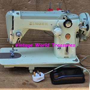 Puede incluir: Máquina de coser Singer vintage en color verde claro. La máquina tiene el logotipo de Singer y varios diales y palancas. Incluye un pedal y un cable de alimentación. La máquina está sobre una superficie de madera.