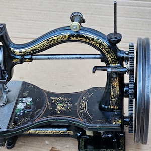 May include: Antique black sewing machine with gold accents and a floral design on the base. The machine has a large hand wheel and a needle bar.