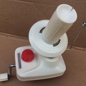 Puede incluir: Enrolla hilo blanco con una perilla roja y una bobina de plástico blanca. El enrollador tiene una manivela y un alambre guía de metal. La etiqueta en el enrollador dice "Speed-Knit Knitmaster".