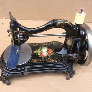 May include: Antique black sewing machine with gold accents and a floral design. The machine has a foot pedal and a spool of thread.