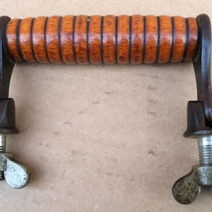 Könnte beinhalten: Ein antiker Holzgriff mit geripptem Design, verbunden mit einem Metallrahmen mit Flügelmuttern. Das Holz hat eine warme braune Farbe und das Metall hat ein rustikales, gealtertes Aussehen. Der Griff stammt wahrscheinlich von einem Vintage-Werkzeug.