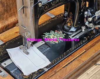 May include: A vintage black sewing machine with a wooden case. The machine has a floral design on the side and the words "SUPERB" and "Vintage World Treasures" are visible. The machine is open and a piece of white fabric is placed under the needle.