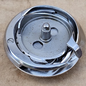 May include: A shiny, silver-colored sewing machine bobbin case. The case has a circular design with a central spindle and various internal components. The metal has a polished finish, reflecting light. The bobbin case is a sewing machine part.