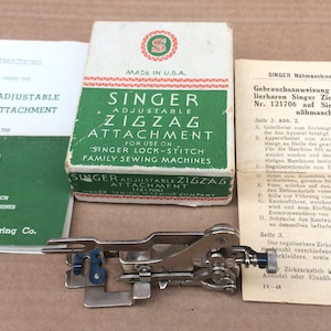 Könnte beinhalten: Ein Vintage Singer verstellbarer Zickzack-Nähmaschinenaufsatz in einer grün-weißen Schachtel mit Anleitung. Die Schachtel ist mit "Singer Adjustable Zigzag Attachment for use on Singer Lock-Stitch Family Sewing Machines" beschriftet.