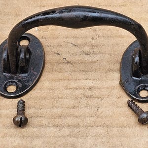 May include: Black metal door handle with two oval-shaped plates and four screws. The handle is curved and has a smooth finish.