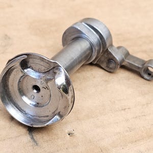 May include: A silver-coloured metallic component, possibly from machinery. It features a circular, concave end, a cylindrical body, and a small articulated arm. The object is lying on a neutral-toned surface, suggesting a mechanical context.