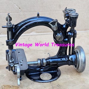 Op de afbeelding: Een vintage zwarte naaimachine met een gebogen arm en een zilveren wiel. De machine heeft ingewikkelde details en een klein cirkelvormig embleem op de basis. De tekst "Vintage World Treasures" is zichtbaar.