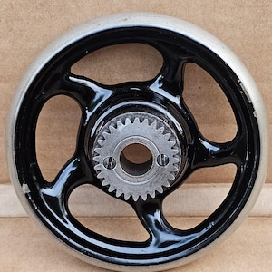 May include: A vintage sewing machine hand wheel with a black, three-spoke design and a silver rim. The center features a gear with a central hole and two screws. The wheel is likely made of metal.