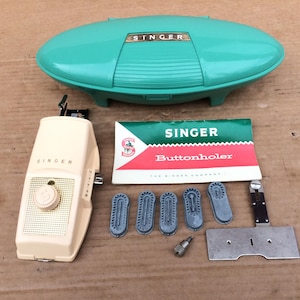 May include: A vintage Singer buttonholer attachment set including a teal oval storage case, a cream-colored sewing machine attachment with a dial, five metal pattern cams, a metal throat plate, a small screw, and an instruction manual.