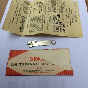 Puede incluir: Un enhebrador de aguja universal Singer, modelo 121632, de color plateado, con instrucciones para enhebrar agujas de máquina de coser. El embalaje es rojo y blanco con el logotipo de Singer y el texto "Enhebrador de aguja universal".