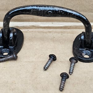 May include: Black metal handle with two oval-shaped mounting plates and four screws. The handle has a curved design and is suitable for a door, drawer, or cabinet.