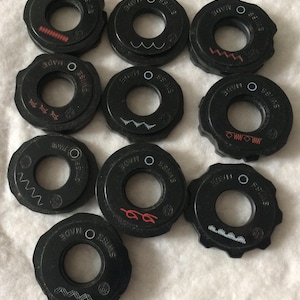 May include: A collection of black sewing machine parts, each with a central hole and the text "MADE SWISS". Some parts feature red or white decorative designs, including wavy lines and zigzags.