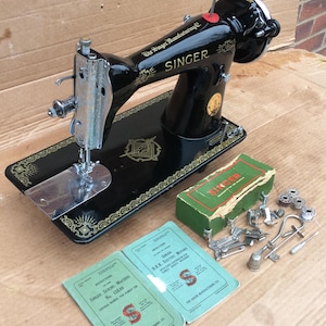 May include: A black Singer sewing machine with a green box and various accessories. The machine has a gold-colored design on the base and the words "The Singer Manufacturing Co." and "SINGER" are printed on the machine.