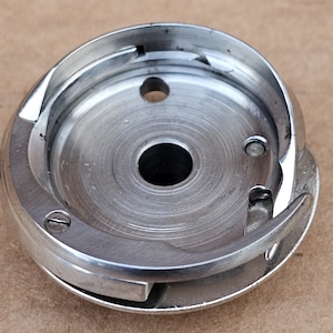 May include: A silver sewing machine bobbin case with a metal hook and a small screw.