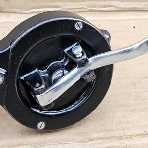 May include: A vintage manual hand-cranked tool with a circular black metal housing, a silver-toned metal arm, and a dark brown wooden handle. The device features exposed screws on the black casing and a small metal mechanism extending from the side.