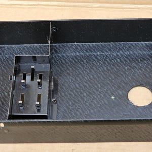 May include: A black rectangular box with a textured surface. It features a metal component with slots and a circular hole. The box is likely a component of a larger device or machine.