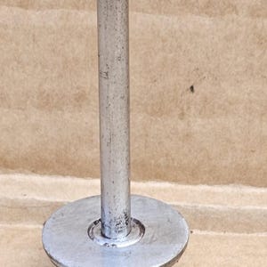 May include: A metal component with a cylindrical rod and a circular base. The rod is topped with a rounded end and is attached to a flat, circular base with a threaded screw. The metal has a worn, used appearance.