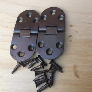 May include: Two antique brown metal hinges with rounded ends and multiple screw holes. A collection of matching screws is scattered below the hinges. The hardware is likely for furniture or a similar woodworking project.