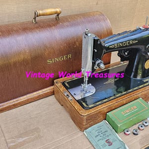 May include: A vintage Singer sewing machine, black with gold detailing, and a wooden base. It is accompanied by a wooden carry case. Accessories include a green box, bobbins, and a manual. The Singer logo is visible.