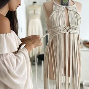 May include: A beige macrame dress on a mannequin, featuring a halter neckline and fringe detailing. A woman is working on the dress, tying the macrame cords. A tarot card is pinned to the mannequin. The dress is a bohemian style.