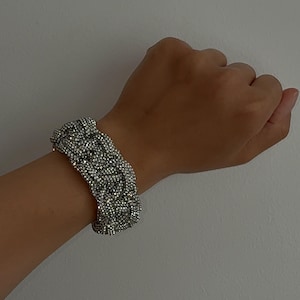May include: A silver-toned bracelet with a braided design, encrusted with numerous small, sparkling crystals. The bracelet is worn on a wrist against a plain, light-colored background. The jewelry piece appears to be a statement accessory.