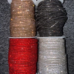 May include: Four spools of rhinestone trim in various colors. The colors include gold, black, red, and silver. Each spool is tightly wound and sits on a white base. The rhinestones sparkle, adding a touch of glamour to the trim.