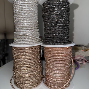 May include: Four spools of rhinestone trim in silver, black, gold, and rose gold. Each spool is tightly wound and sits on a white plastic base. The trim is made of small, sparkling rhinestones, ideal for embellishing clothing or accessories.