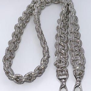Könnte beinhalten: Ein silberner, geflochtener Handtaschenriemen mit funkelnden Strasssteinen. Der Riemen hat zwei silberfarbene Metallverschlüsse zum einfachen Anbringen. Das geflochtene Design verleiht dem Accessoire Textur und visuelles Interesse.