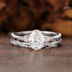 May include: A silver engagement ring set with an oval-cut central stone and smaller accent stones. The band has a twisted design with additional small stones. The ring is displayed on a wooden surface.