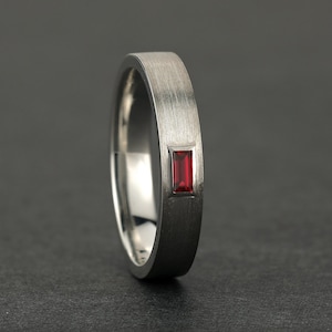 Peut inclure: Une bague en métal argenté avec une finition mate brossée, ornée d'une unique pierre précieuse rouge de taille baguette rectangulaire, sertie à fleur au centre de l'anneau. La bague présente un profil plat et une surface intérieure polie.