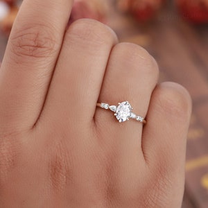 May include: A delicate gold engagement ring with an oval-shaped center stone and smaller accent stones. The ring is set on a thin band.