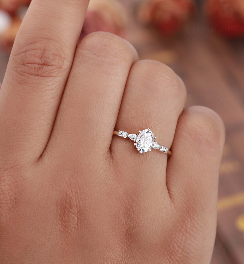 May include: A delicate gold engagement ring with an oval-shaped center stone and smaller accent stones. The ring is set on a thin band.
