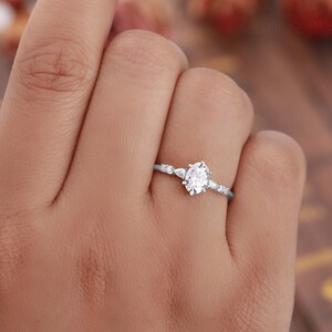 May include: A delicate silver ring with a round, clear center stone and smaller stones on the band. The ring is on a person's finger.