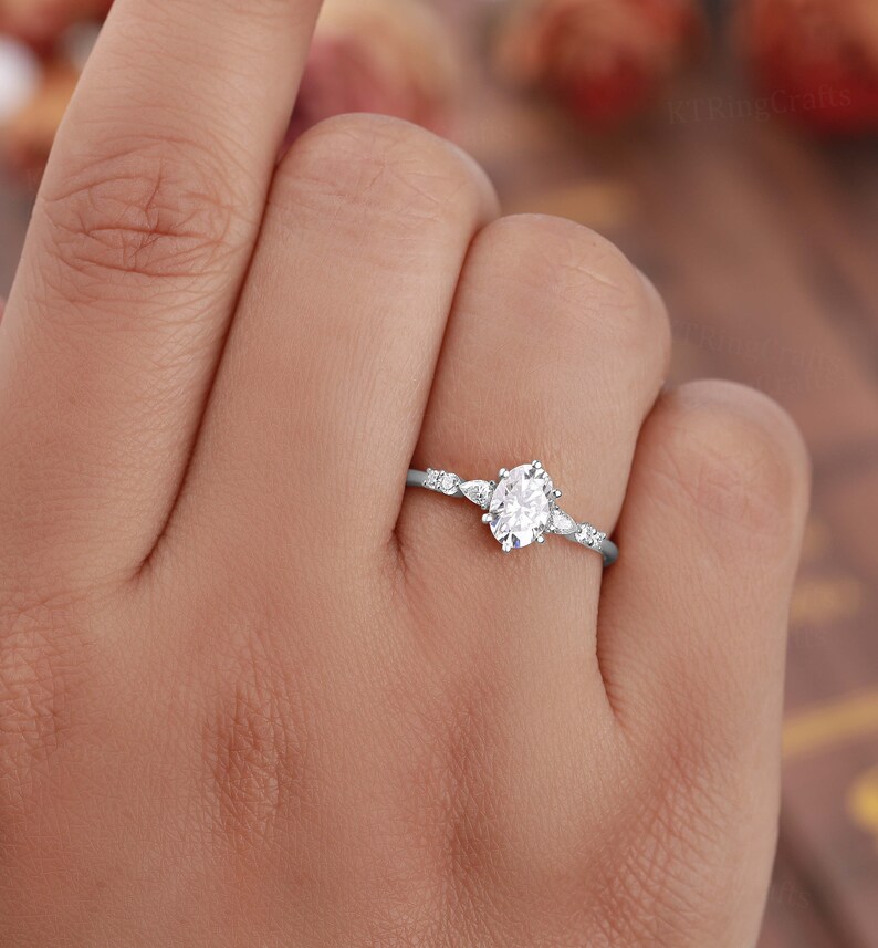 May include: A delicate silver ring with a round, clear center stone and smaller stones on the band. The ring is on a person's finger.
