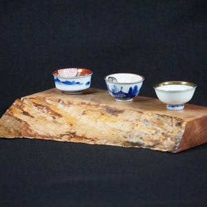 May include: Three small, decorative ceramic bowls with intricate designs rest on a rustic wooden slab. The bowls feature blue, red, and gold accents. The wooden slab has a natural, textured appearance, set against a dark background.