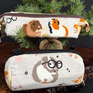 May include: Two cylindrical fabric pencil cases featuring light beige backgrounds with printed guinea pig illustrations. The top case shows two guinea pigs, while the bottom case features a single guinea pig wearing black round glasses.
