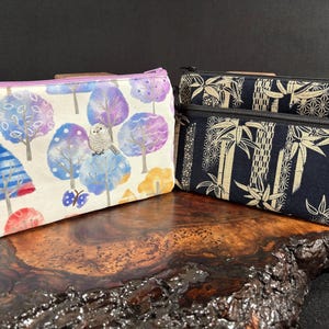 May include: Two zippered pouches. One features a watercolor design of trees in blue, purple, and yellow, with an owl and butterfly. The other two pouches have a navy blue background with a cream-colored bamboo pattern. The pouches are on a dark wooden surface.