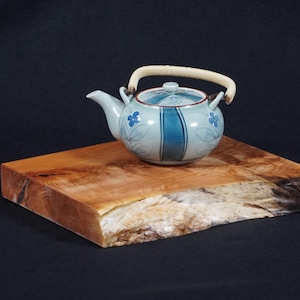 May include: A light blue and white ceramic teapot with a floral design and a cream-coloured handle sits on a rectangular wooden block. The teapot has blue stripes and floral accents. The wooden block is approximately 30 cm long.
