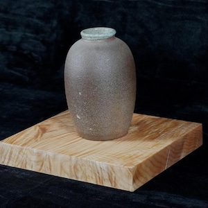 May include: A small, brown ceramic vase with a light green glaze on the rim, sitting on a wooden base.