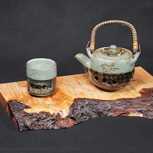 May include: A tea set comprising a ceramic teapot with a woven handle and a matching cup, in green and brown tones. The set is displayed on a wooden serving board with a natural edge, highlighting the wood grain.