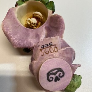 May include: A purple and green ceramic flower figurine with a small figure inside. The flower has a circular design on the bottom with the number 3600 and a swirl design.