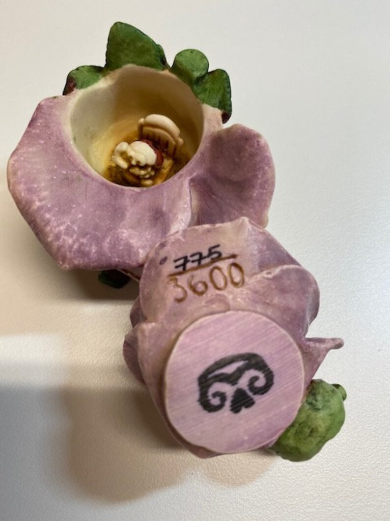 May include: A purple and green ceramic flower figurine with a small figure inside. The flower has a circular design on the bottom with the number 3600 and a swirl design.