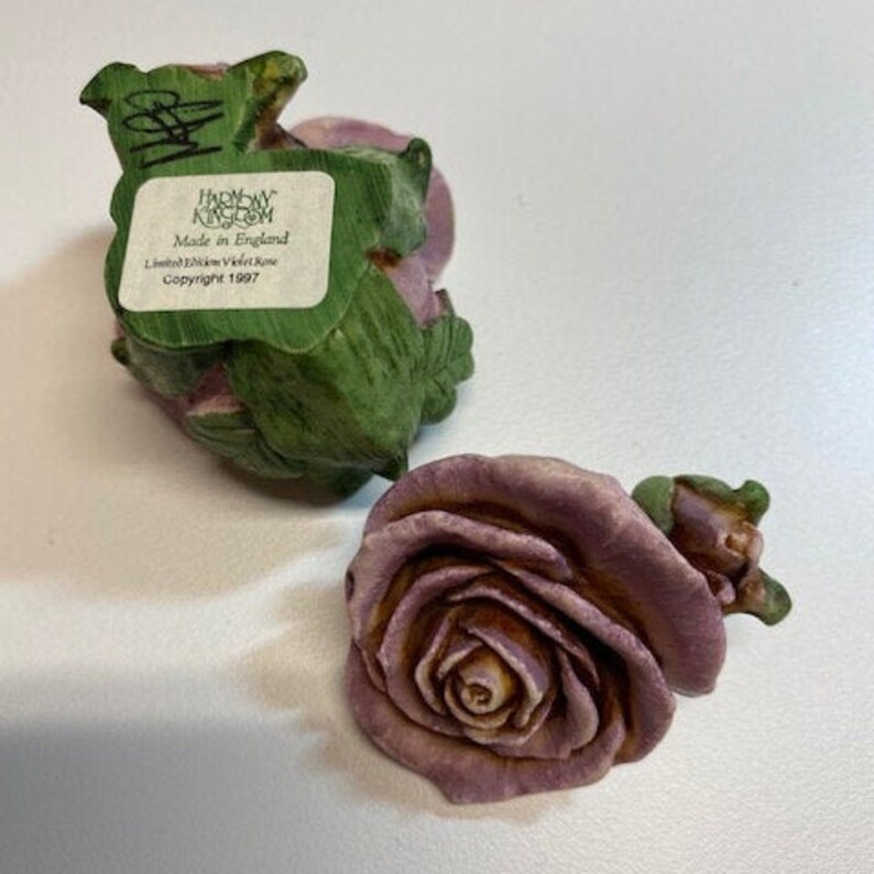 May include: Two small ceramic figurines of purple roses. One rose is green and has a label that reads "Harmony Kingdom Made in England Limited Edition Violet Rose Copyright 1997".