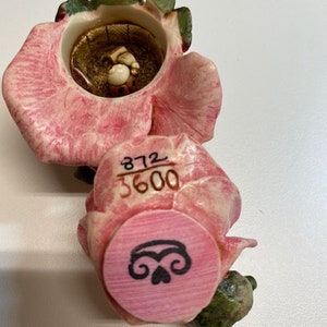 May include: A pink and green ceramic rose figurine with a small, open box inside. The box has a small, white figure inside. The bottom of the figurine has a pink circle with a black design and the numbers "372 3600" printed on the side.