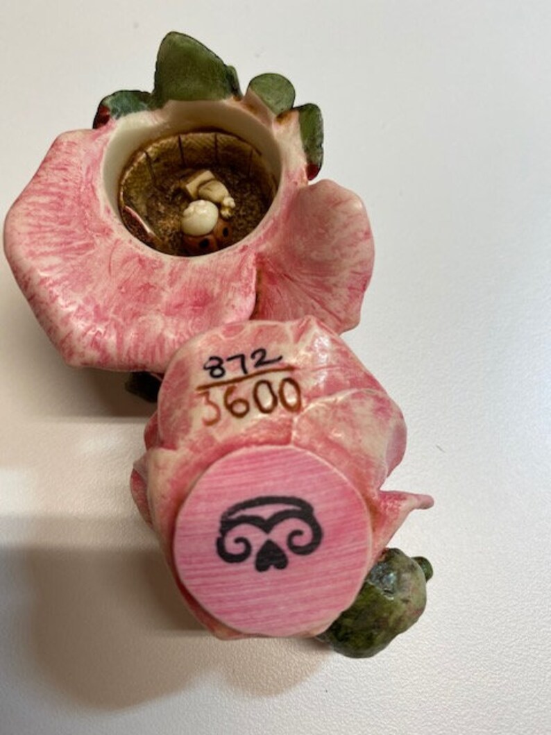 May include: A pink and green ceramic rose figurine with a small, open box inside. The box has a small, white figure inside. The bottom of the figurine has a pink circle with a black design and the numbers "372 3600" printed on the side.