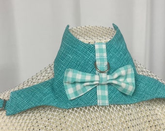 May include: A teal blue dog harness with a white and teal gingham bow tie. The harness has a metal D-ring and a buckle closure.