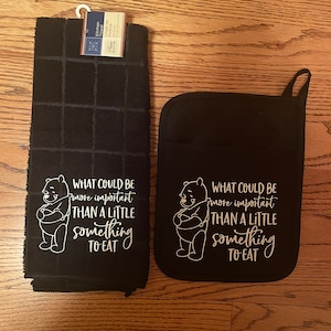 Puede incluir: Juego de paño de cocina y agarradera negros con un gráfico de Winnie the Pooh y el texto "WHAT COULD BE more important THAN A LITTLE something TO EAT". La toalla tiene un patrón de cuadrícula.